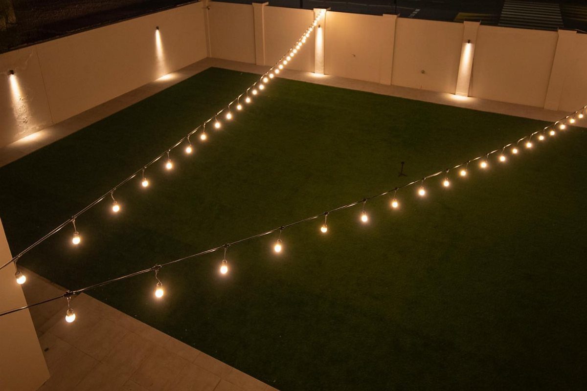Strings of warm glowing light bulbs hanging over a dark green backyard lawn.