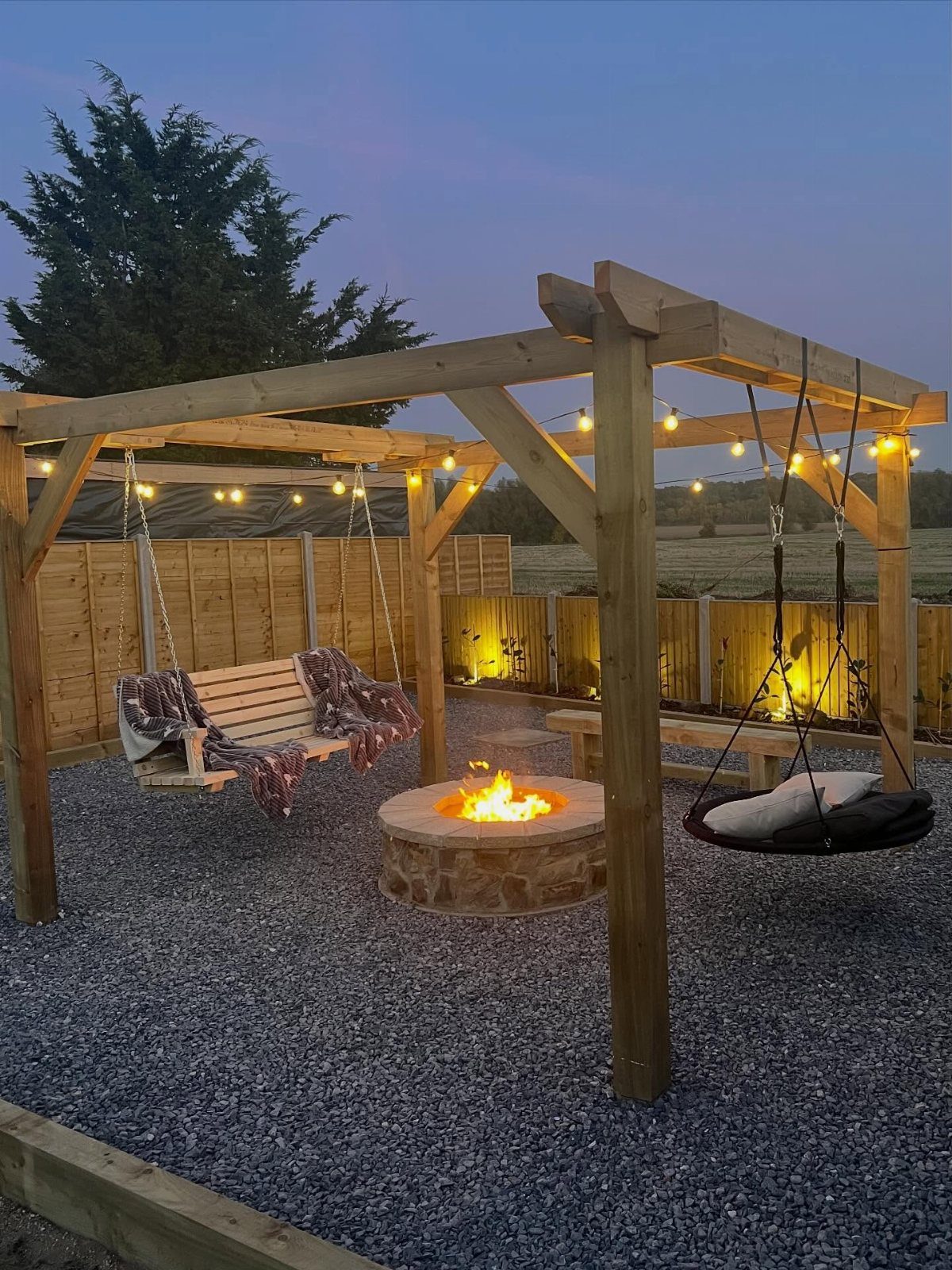 A stone fire pit with flames sits under a wooden pergola with hanging swings and string lights.