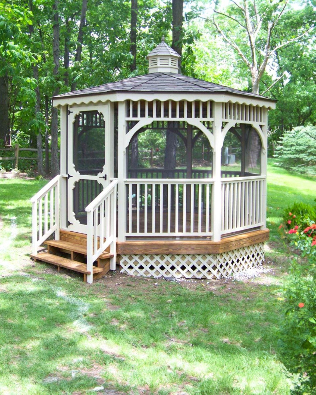 A white screened-in wooden gazebo with a dark roof stands on a grassy lawn near trees.