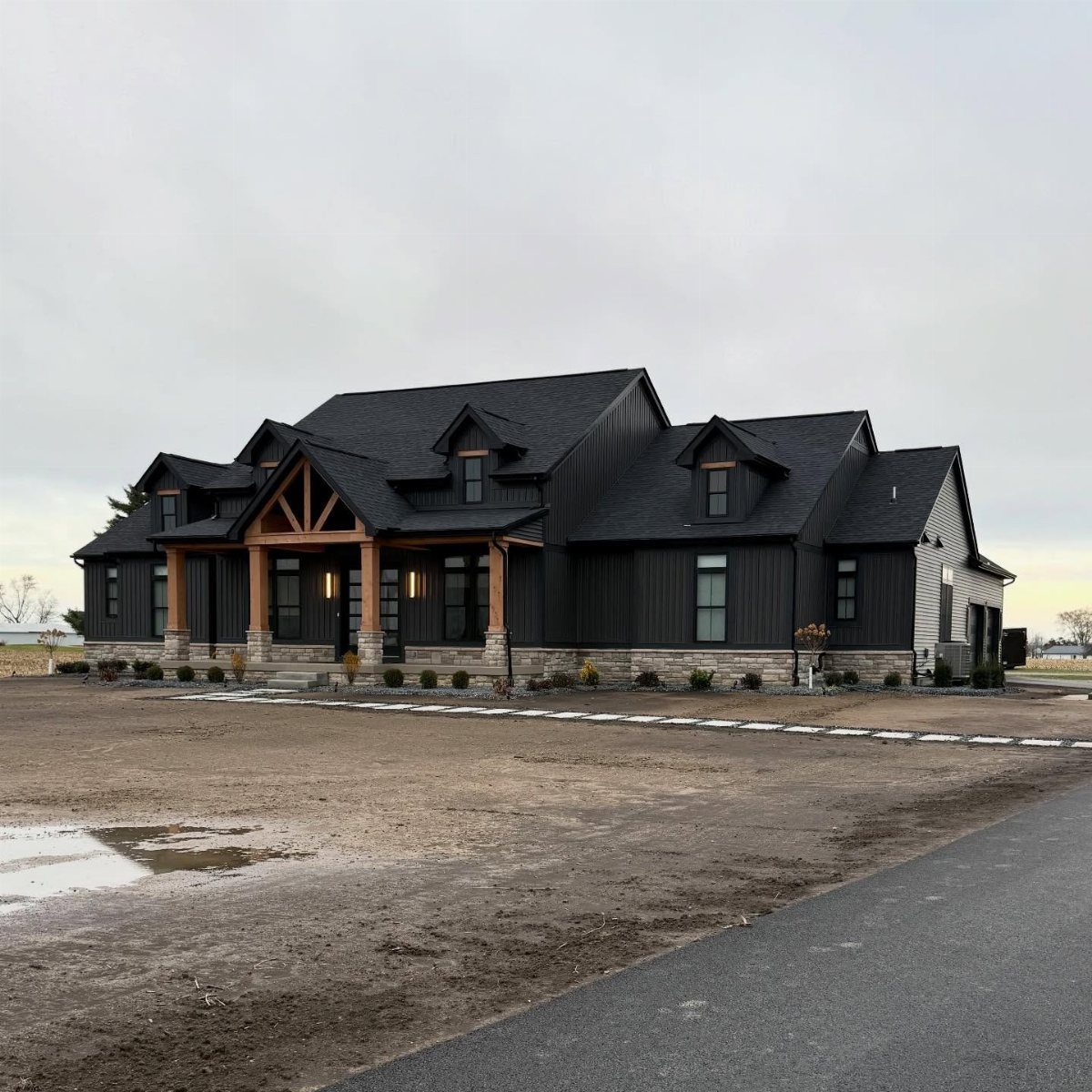 Black modern farmhouse with wooden porch accents and stone base situated on a dirt lot.