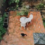 Reddish-orange brick patio with white wrought-iron garden furniture and a black and white cat.