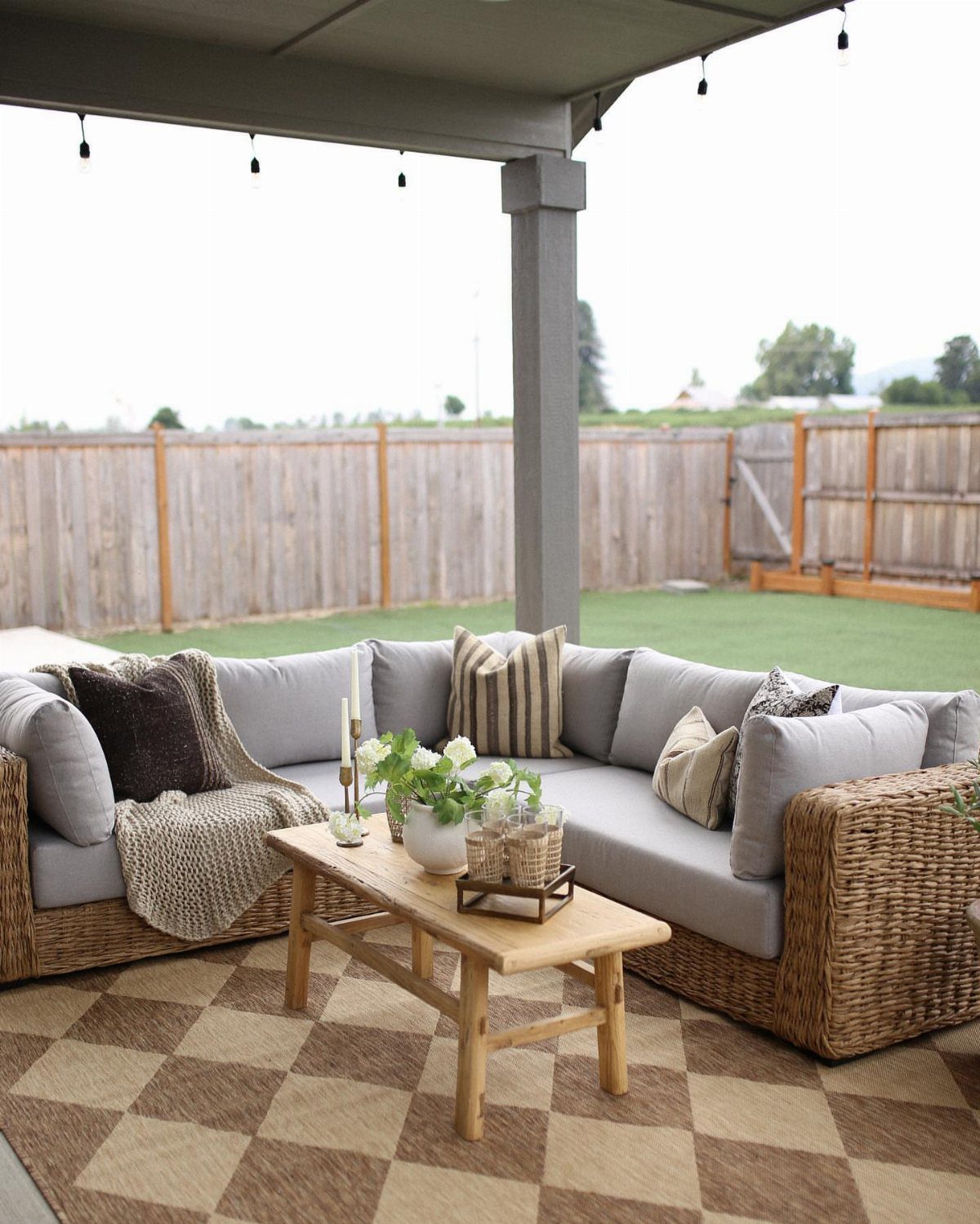 Grey outdoor sectional sofa with cushions and a wicker base on a checkered rug.