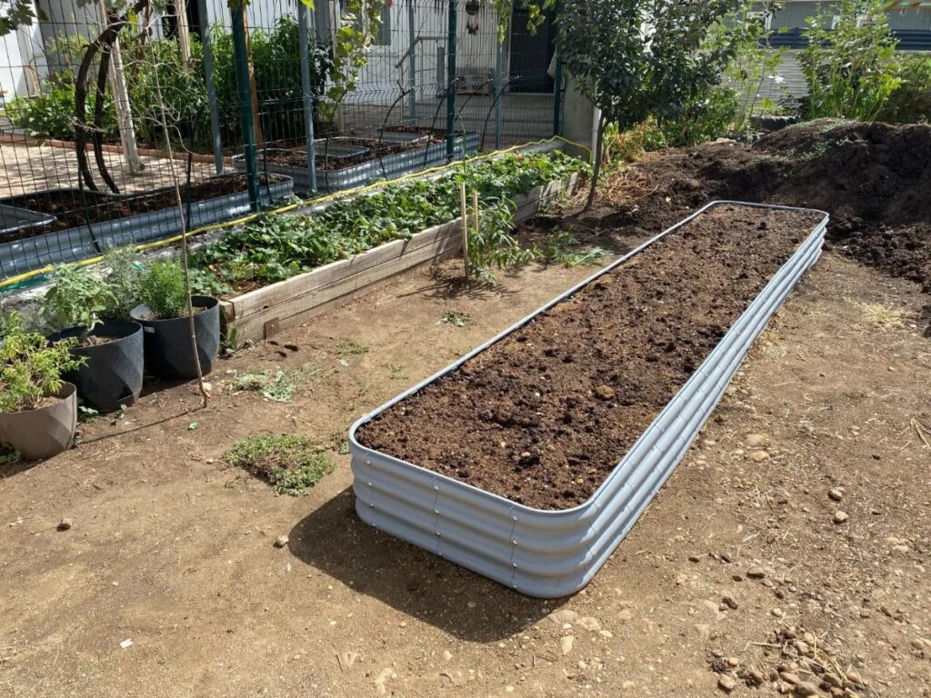 A long corrugated metal raised garden bed filled with dark soil in a backyard garden.